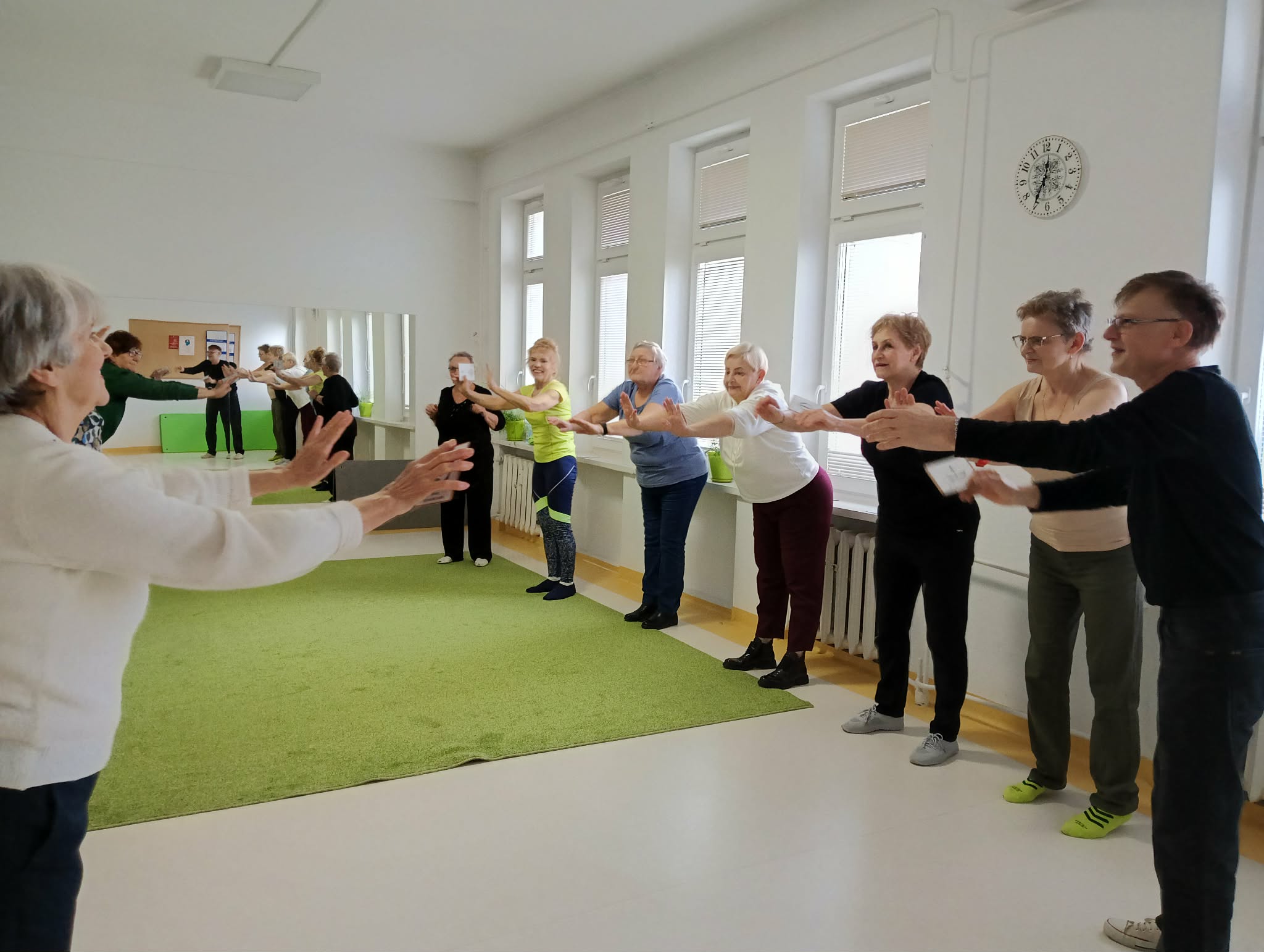 Grupa seniorek i seniorów stoi w kręgu w jasnej sali z wyciągniętymi przed siebie rękami.
